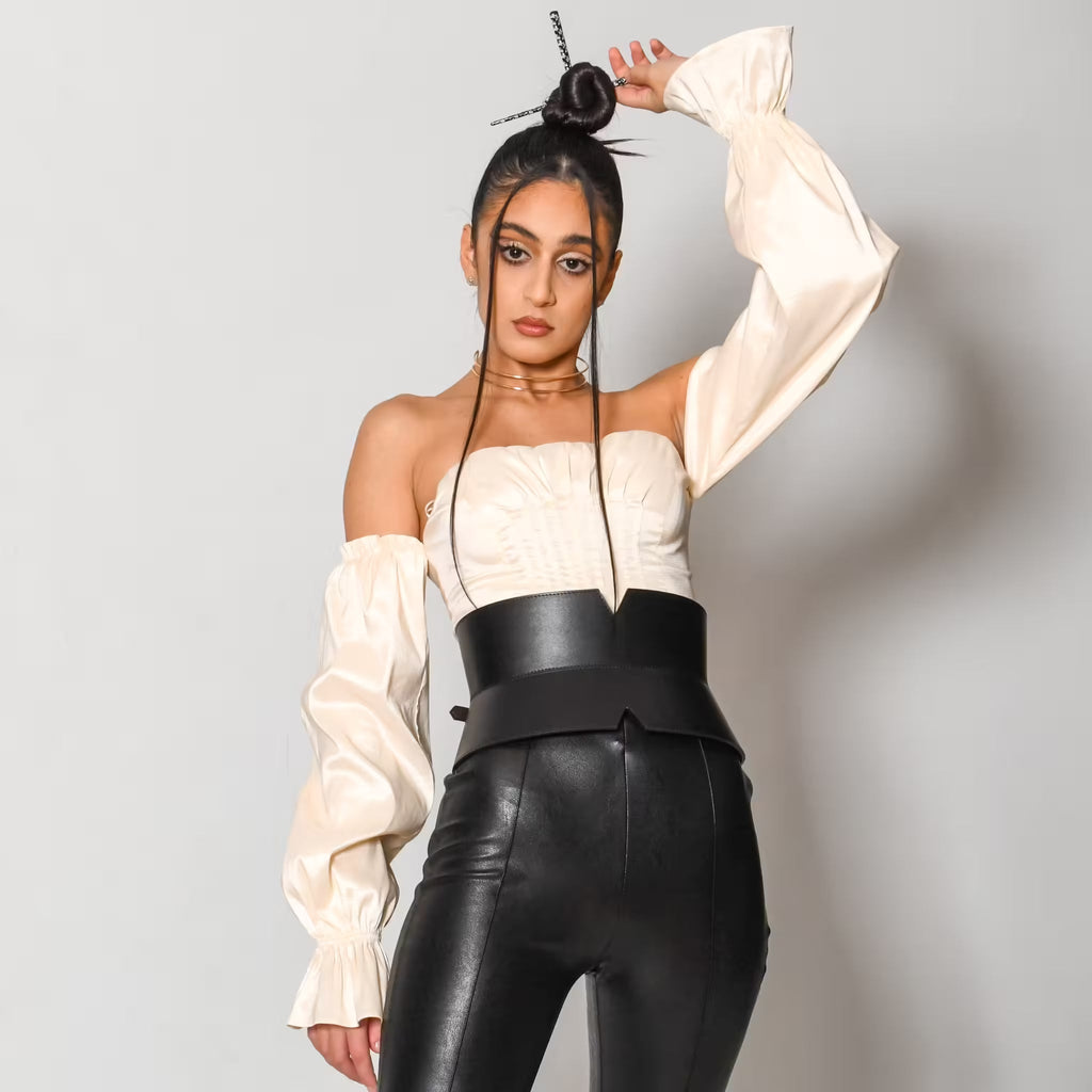 Aria Margo black leather V-cut corset belt worn over a cream off-shoulder top and black leather trousers.