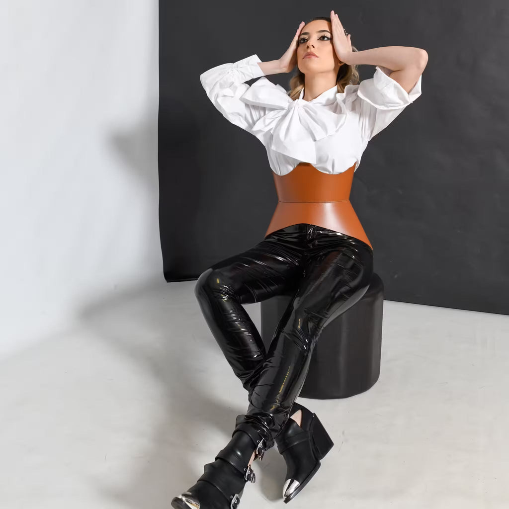 Aria Margo brown Italian leather corset belt worn over a white blouse and black trousers by a seated model.
