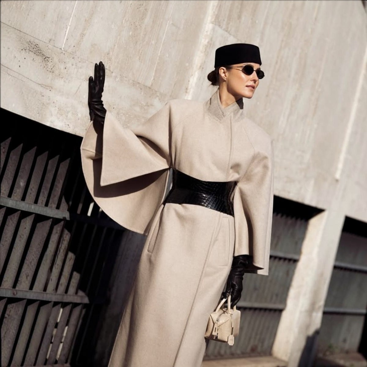 Aria Margo black croc-embossed leather hourglass corset belt worn over a beige wool cape coat with black gloves.