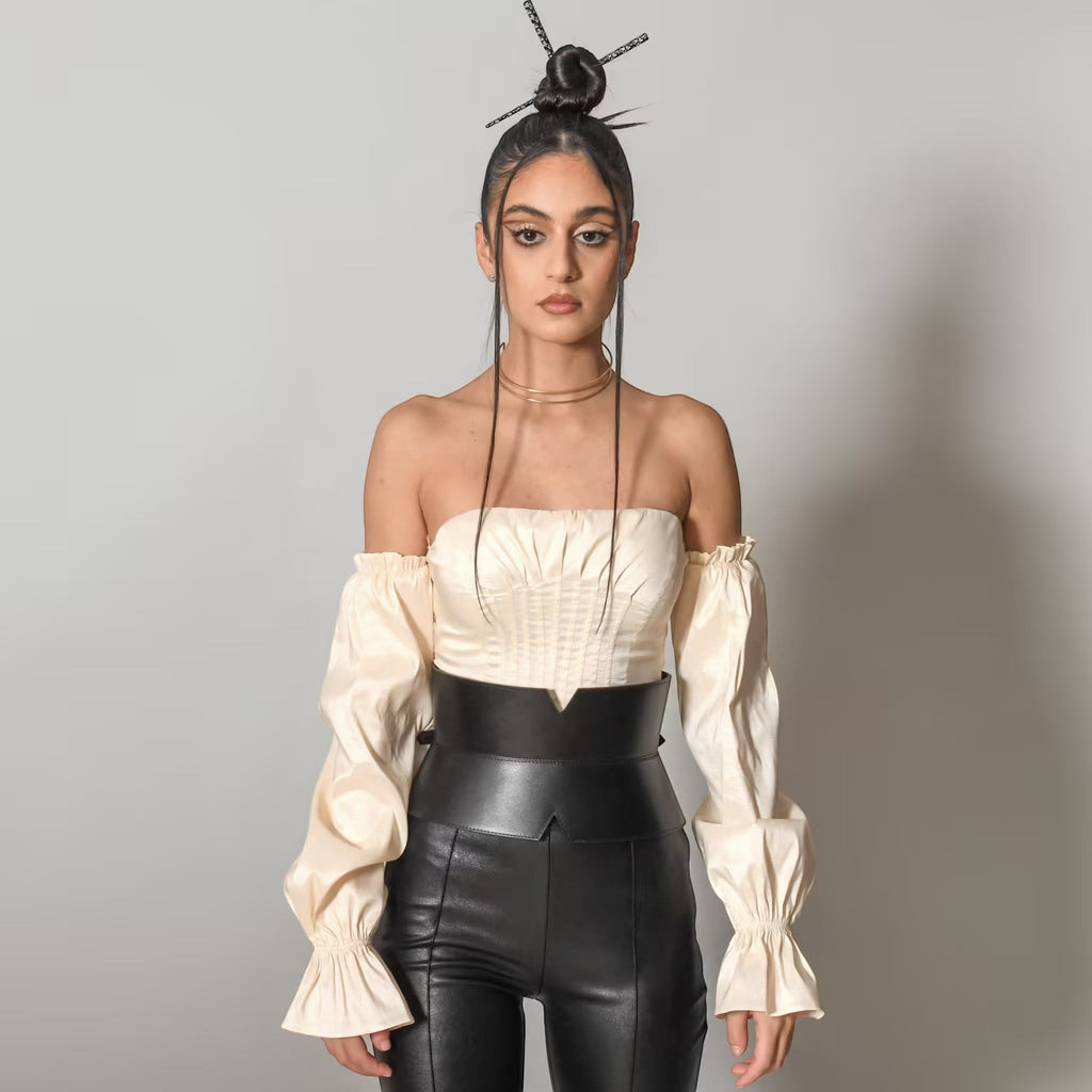 Aria Margo black leather V-cut corset belt worn over a cream off-shoulder top and black leather trousers.