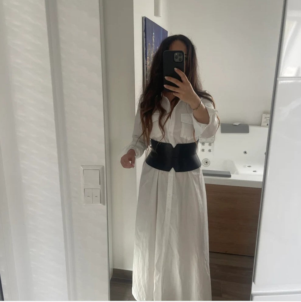 Aria Margo black leather V-cut corset belt worn over a long white button-down shirt dress in a mirror selfie.