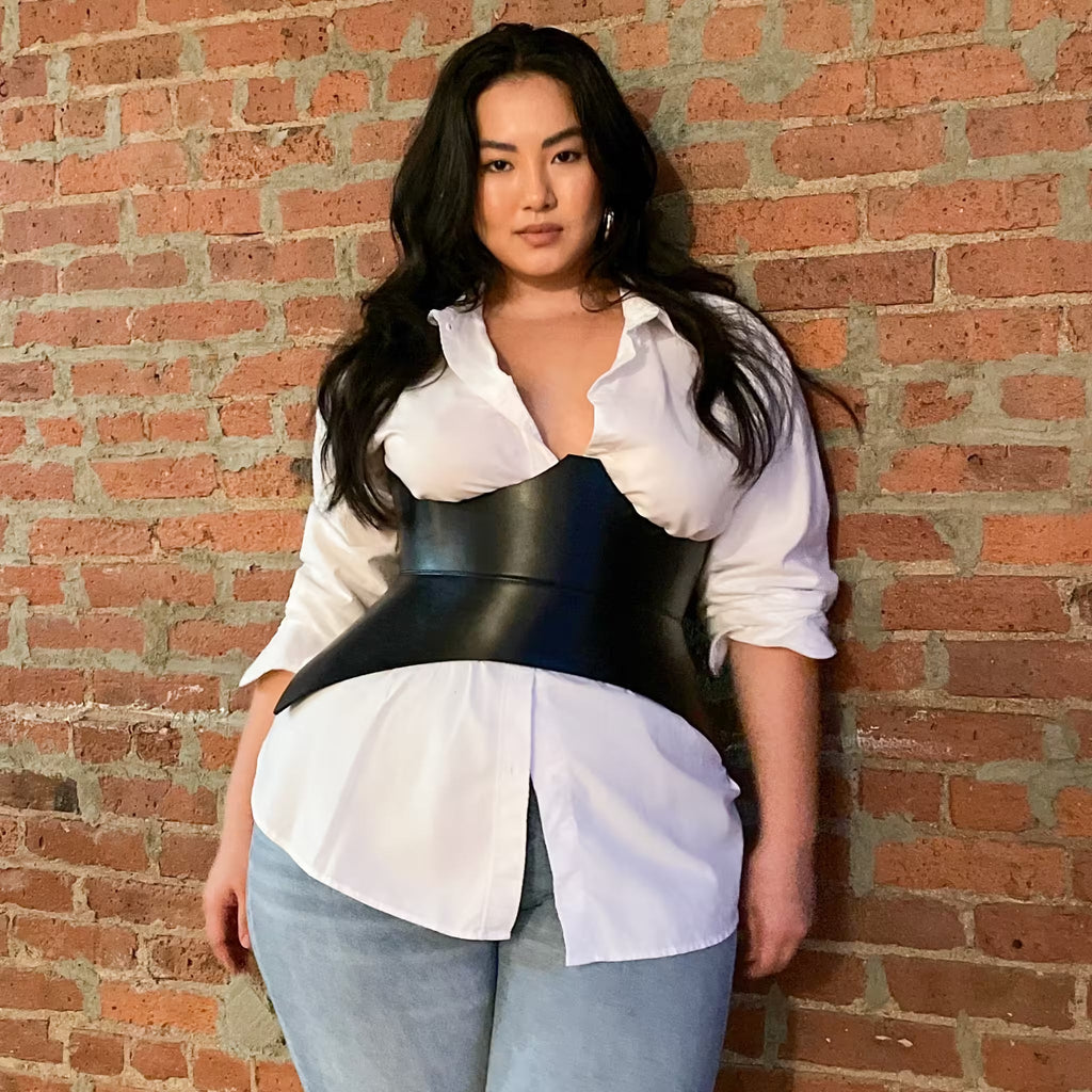 Aria Margo black Italian leather corset belt worn over a white button-down shirt and jeans against a brick wall.