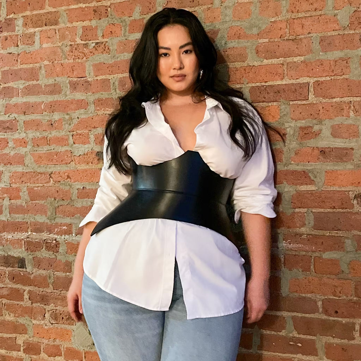 Aria Margo black Italian leather corset belt worn over a white button-down shirt and jeans against a brick wall.