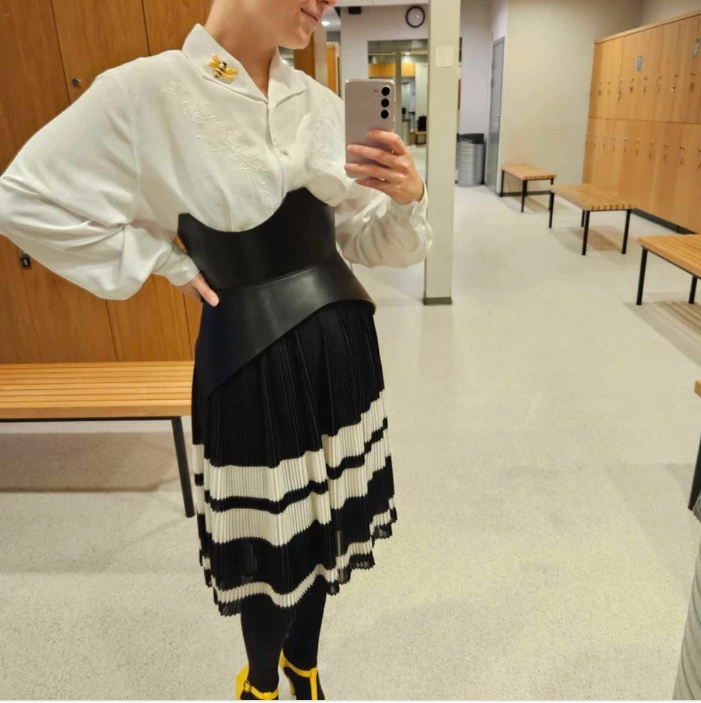Aria Margo black leather corset belt worn over a white shirt and striped pleated skirt for a feminine silhouette