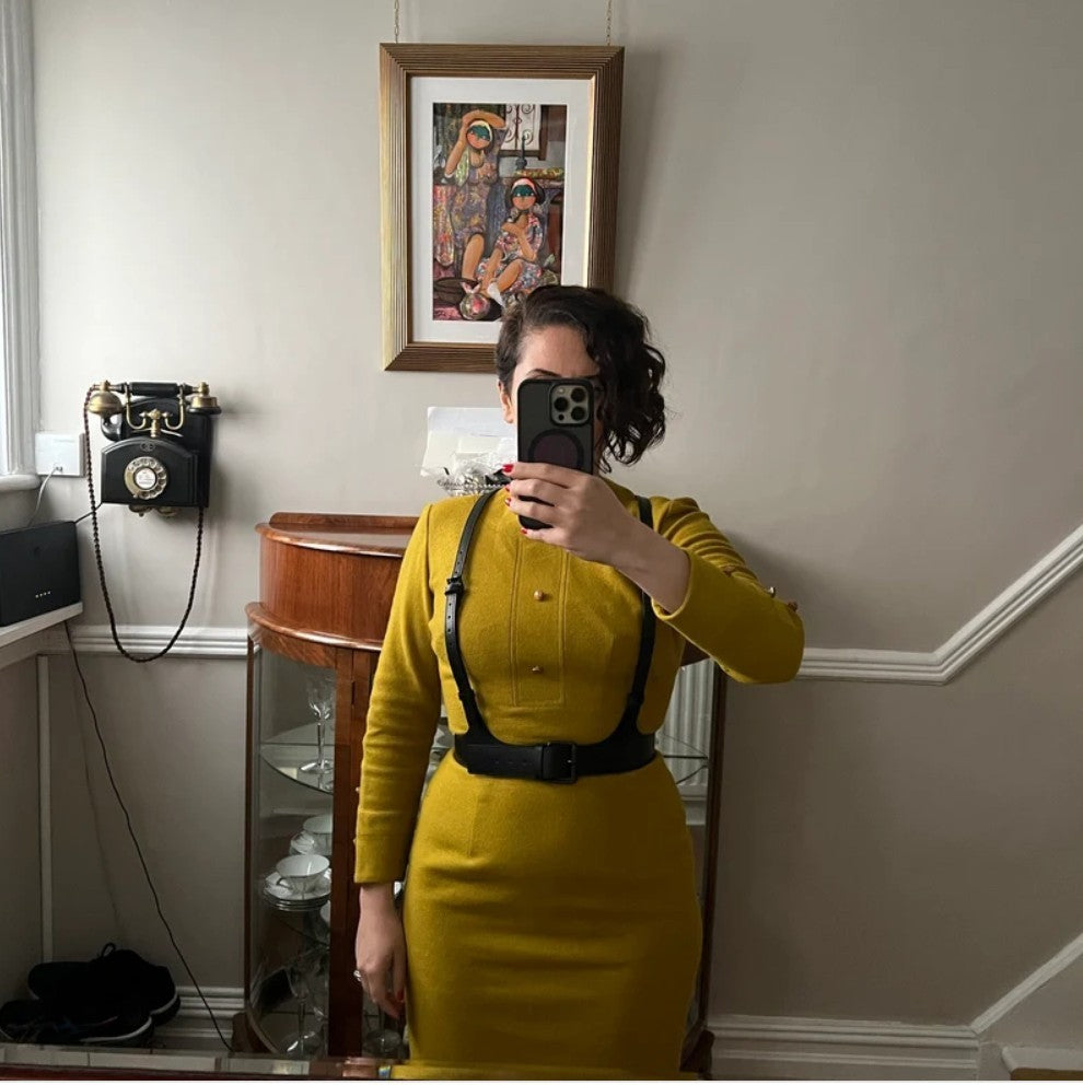 Aria Margo black leather V-neck harness belt worn over a mustard yellow long-sleeved dress in a home interior.