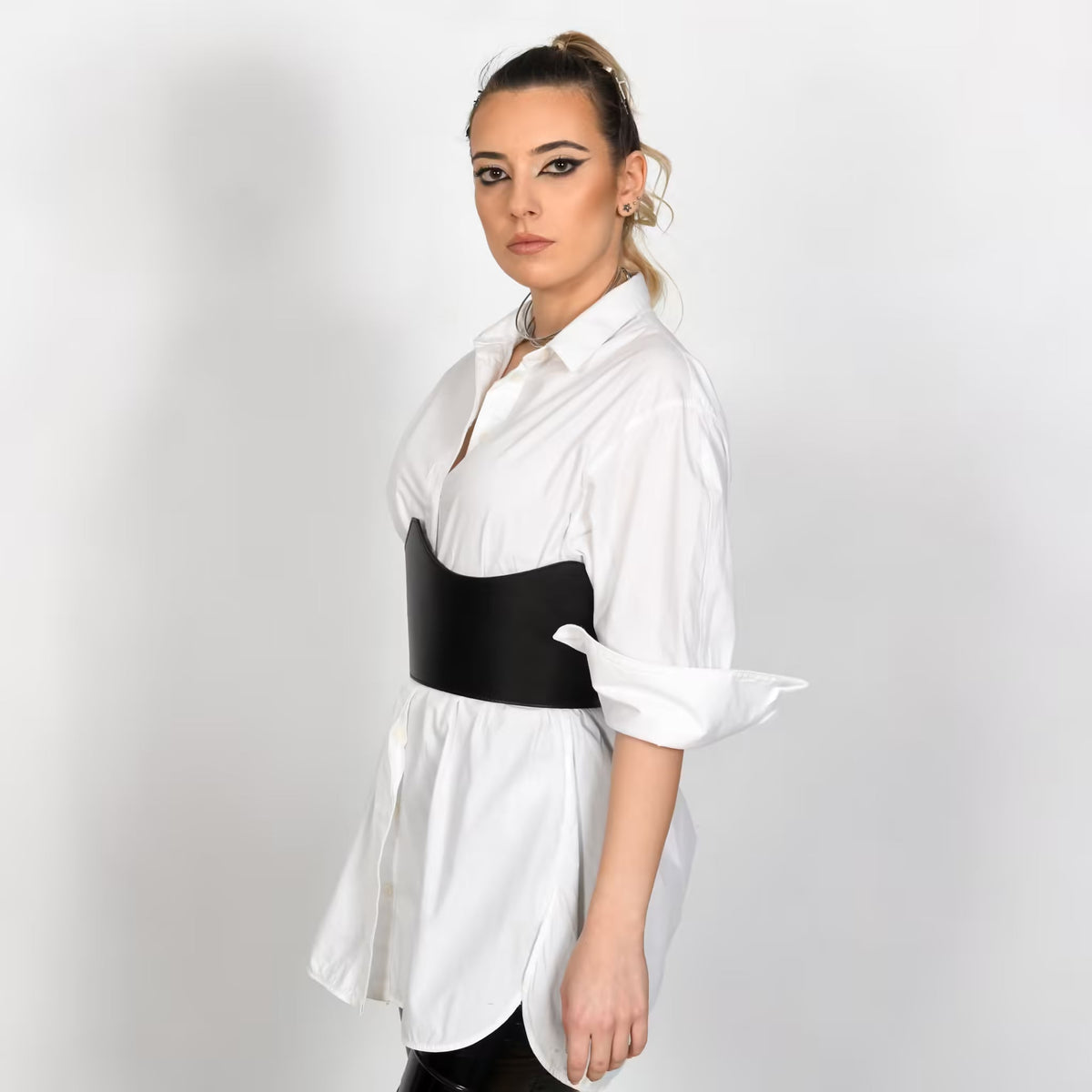 Aria Margo black leather under-bust belt worn over a white button-down shirt by a woman with a high ponytail.