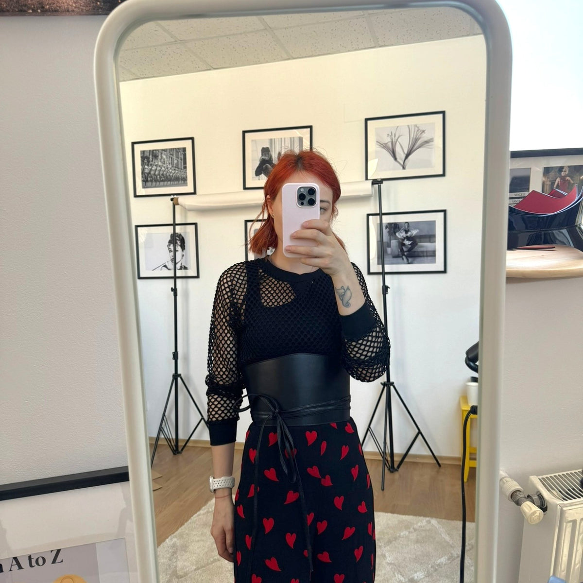 Aria Margo black leather under-bust belt worn over a black mesh top and heart-print skirt in a mirror selfie.