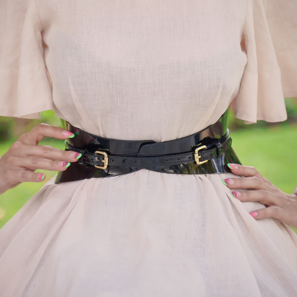 Aria Margo slim black patent leather corset belt worn over a light beige dress to define the waistline.