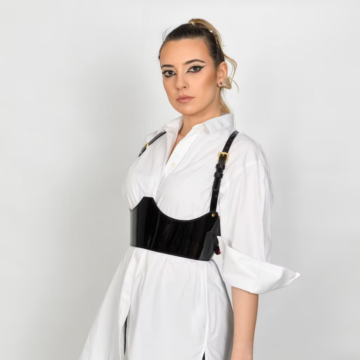 Aria Margo black patent leather under-bust belt with adjustable shoulder straps worn over a white shirt.