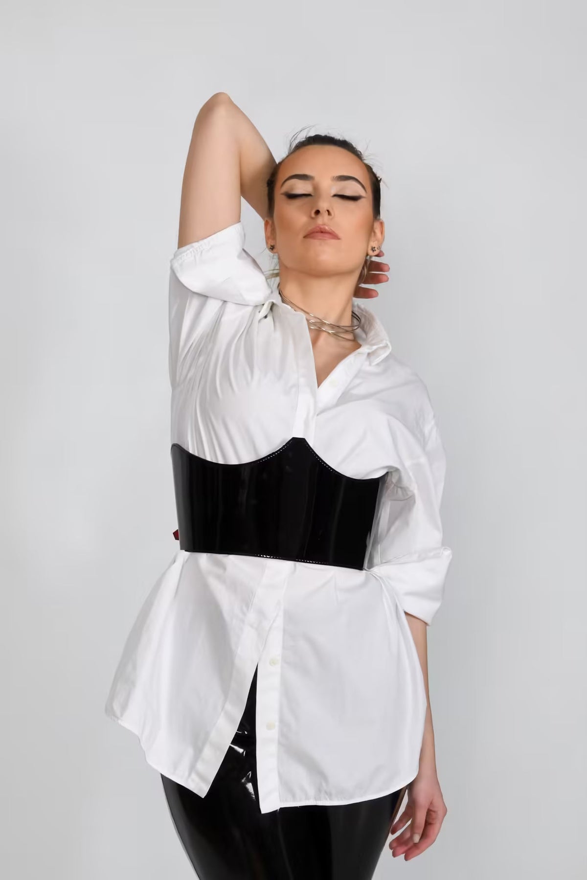 Aria Margo  black patent leather under-bust belt worn over a white button-down shirt for a structured silhouette.