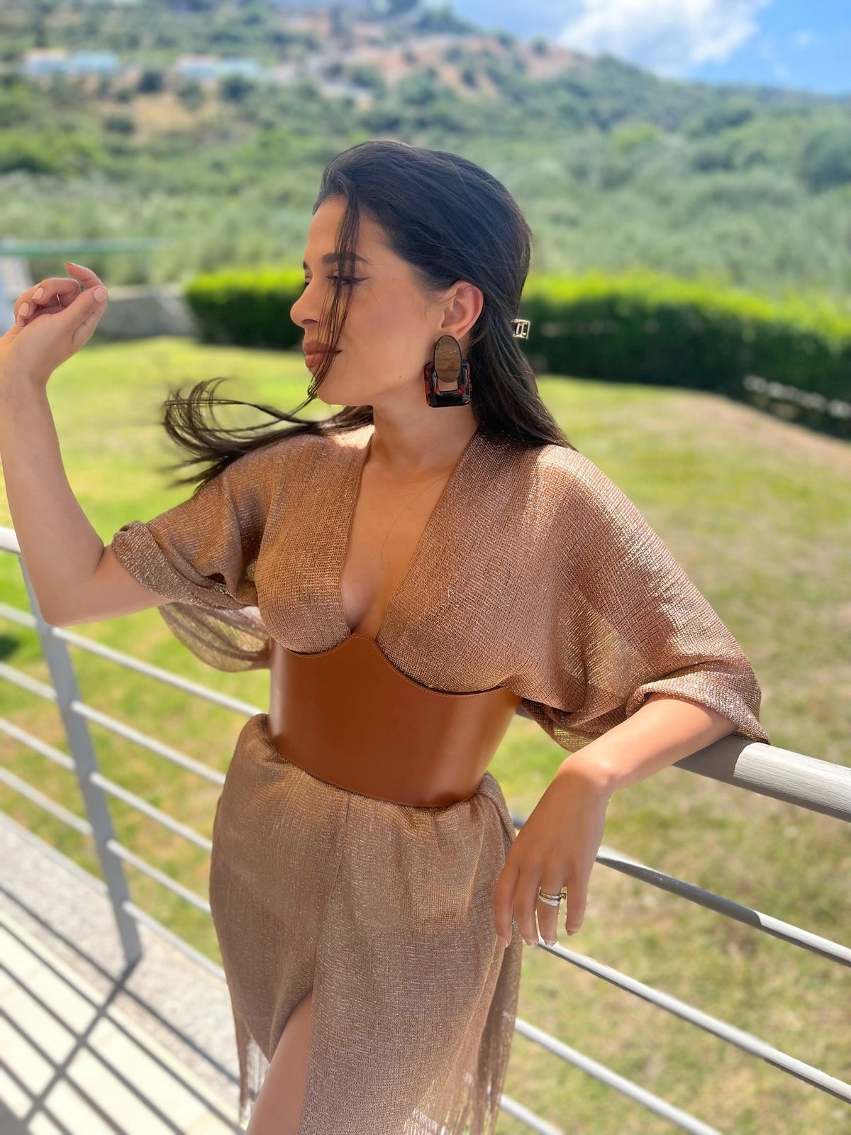 Aria Margo brown leather under-bust belt worn over a shimmering gold dress in an outdoor garden setting.