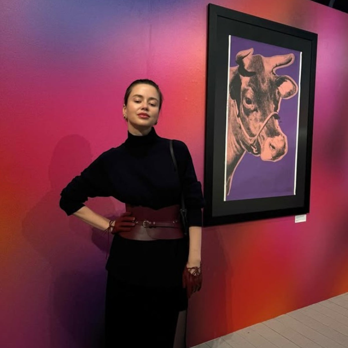 Aria Margo burgundy leather hourglass corset belt worn over a black turtleneck dress in an art gallery.