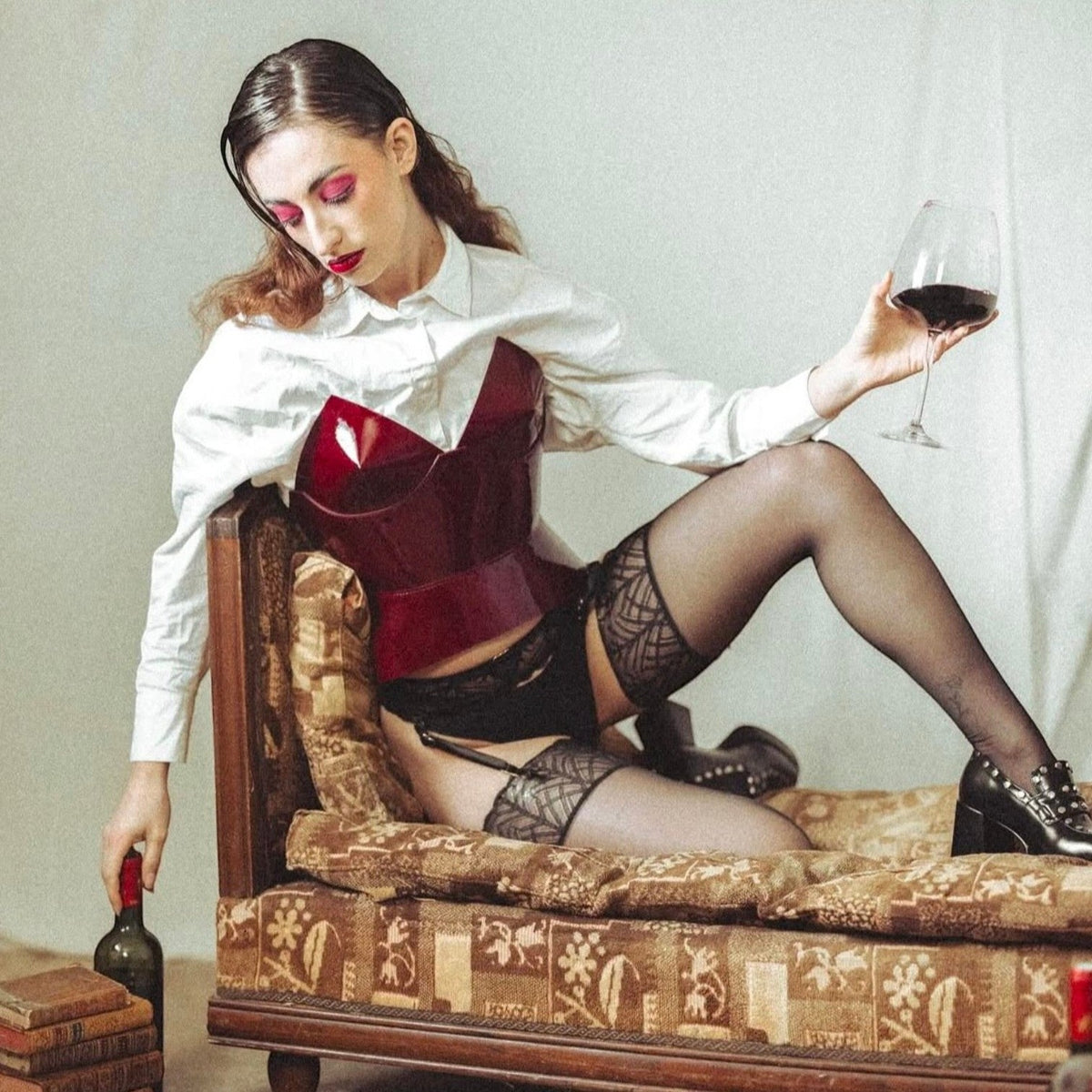 Aria Margo burgundy patent leather corset worn over a white shirt by a woman lounging on a vintage sofa.