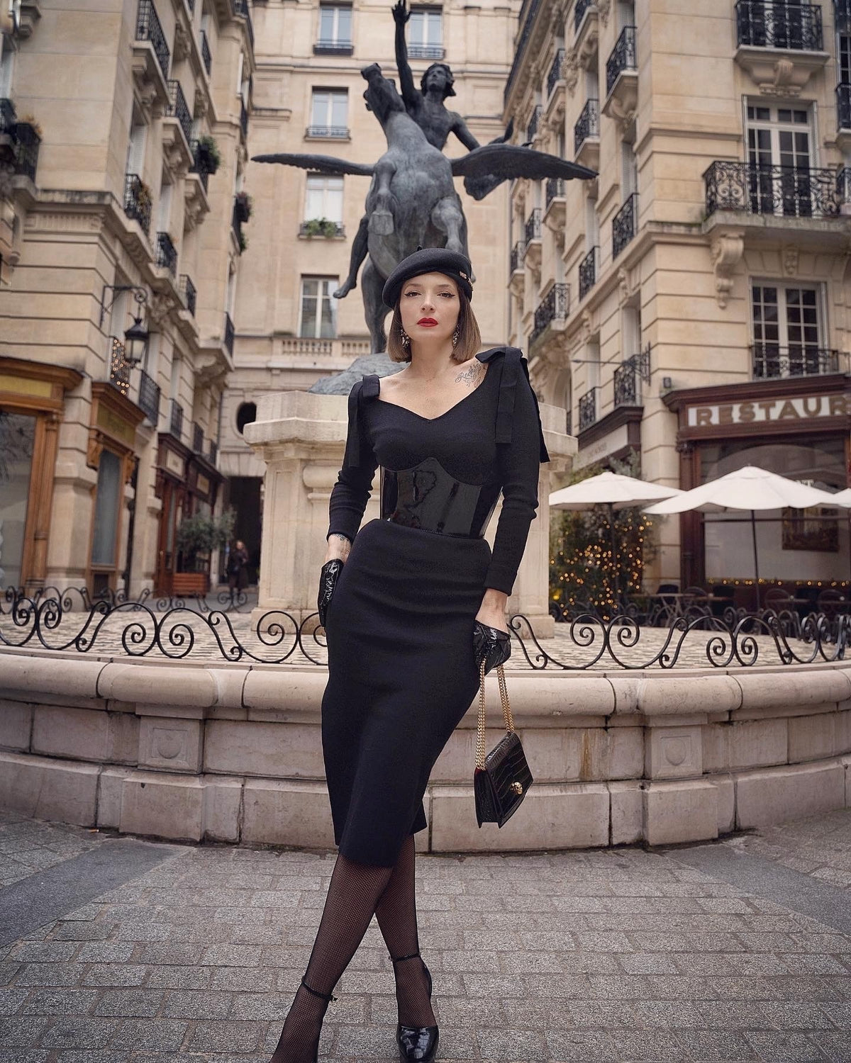 Aria Margo black patent leather under-bust belt worn by a woman in a chic black outfit in a Parisian square.