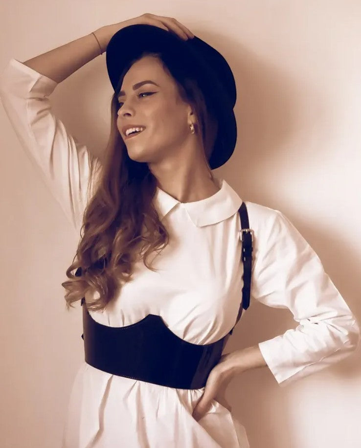 Aria Margo black patent leather under-bust belt with adjustable straps worn over a white collared dress and hat.