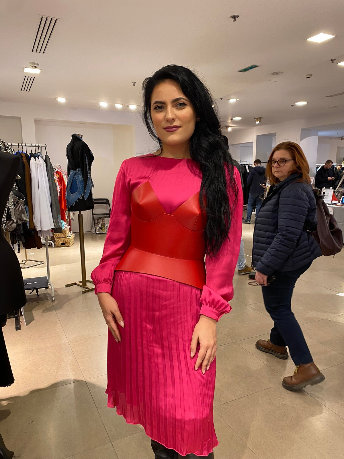 Aria Margo red leather corset belt worn over a long-sleeved pink pleated dress in a retail store setting.