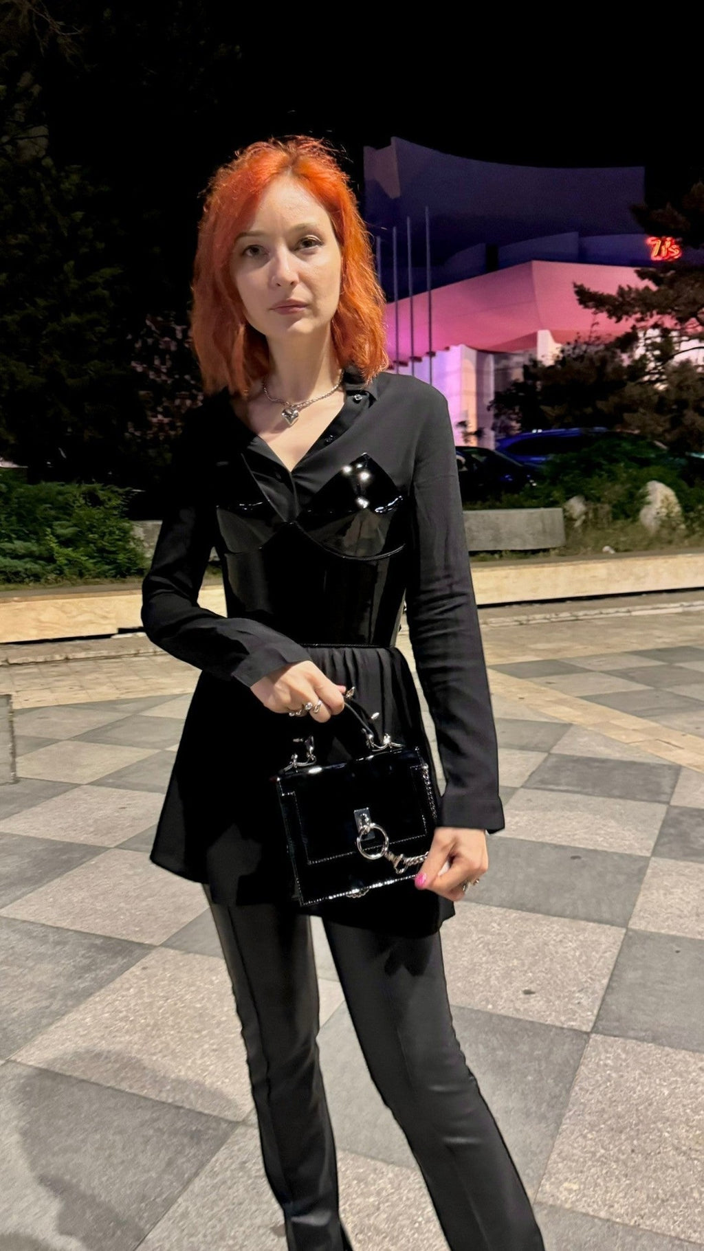 Aria Margo Test black patent leather over-bust corset worn over a black shirt by a woman with red hair at night.