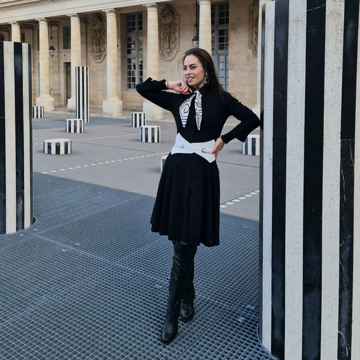 Aria Margo white leather Thorns corset belt worn over a black dress by a woman in a Parisian courtyard.