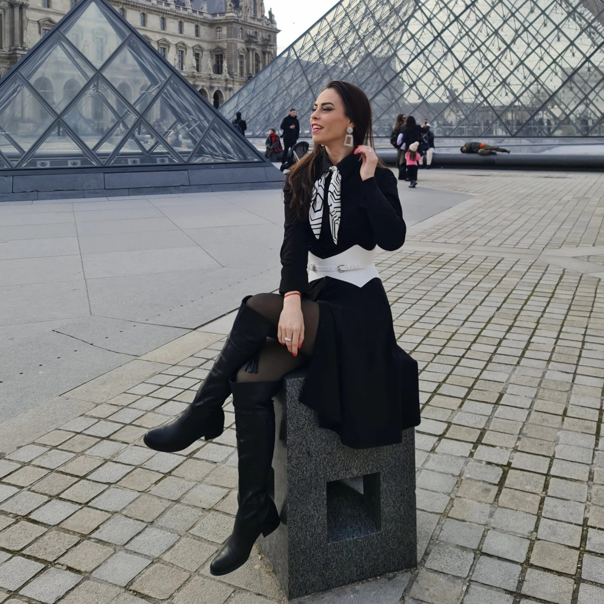 Aria Margo white leather corset belt worn over a black dress by a woman sitting in front of the Louvre pyramid